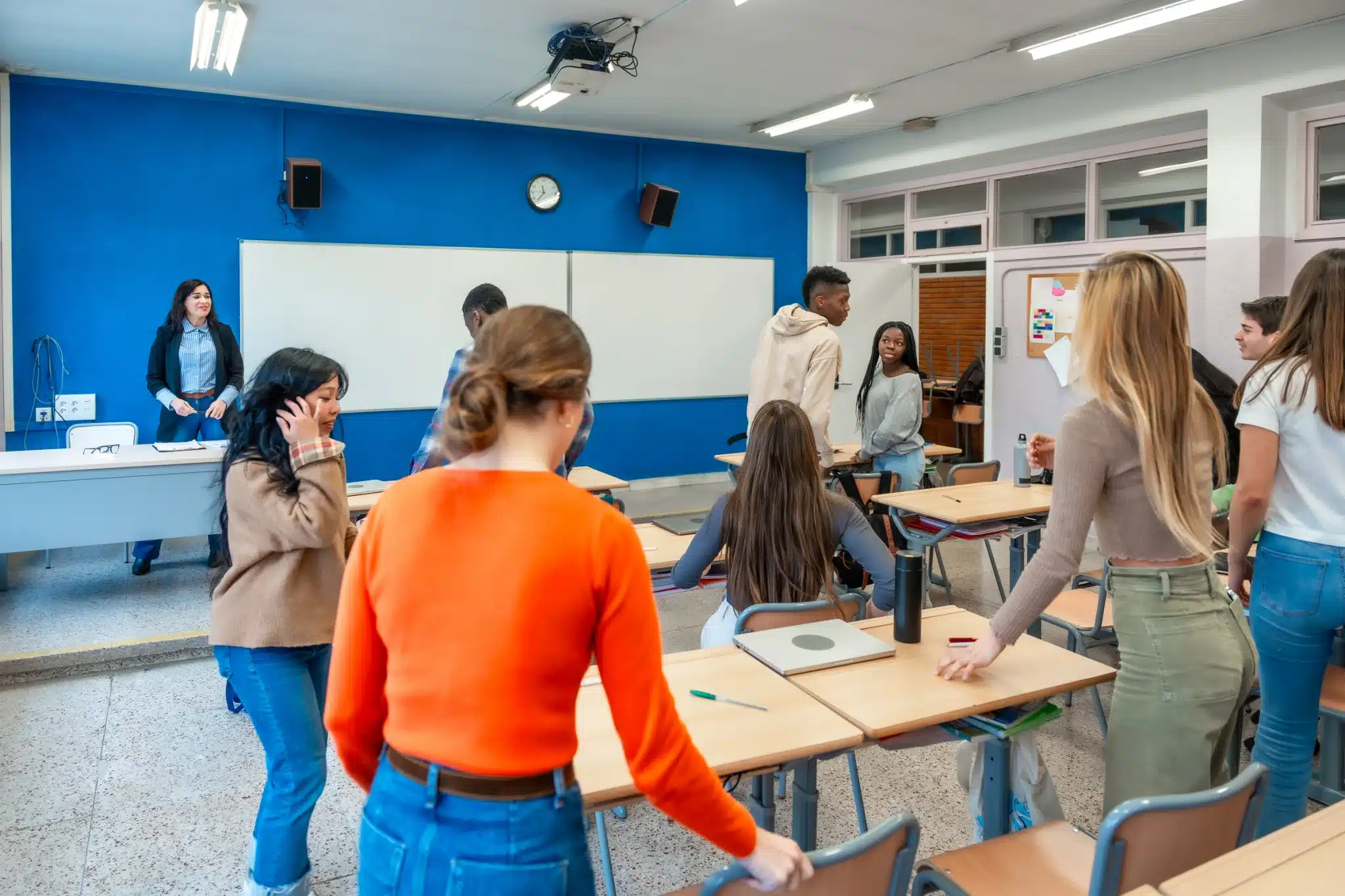 People standing and sitting among desks in a classroom with a blue wall