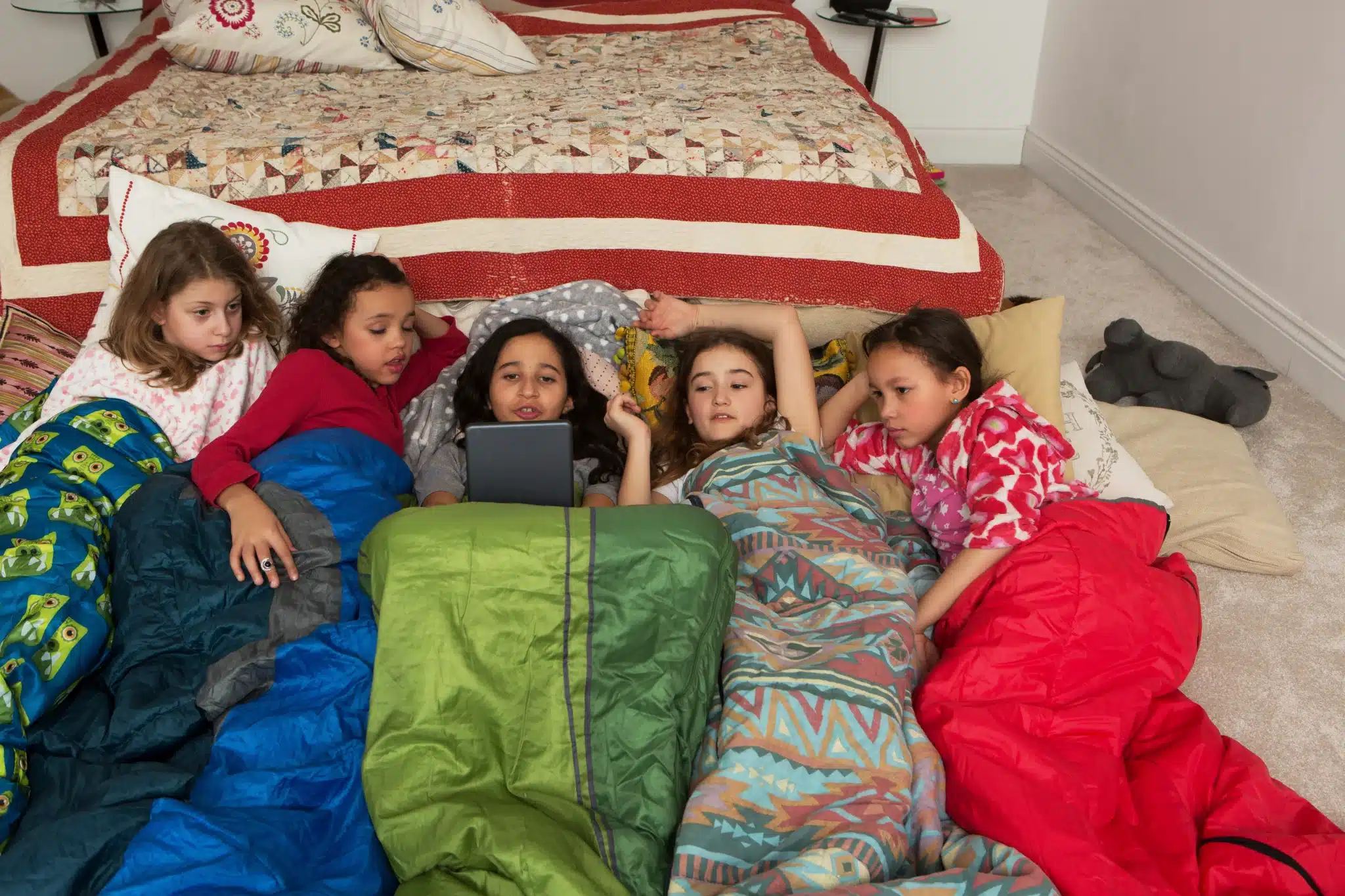 Children in colorful sleeping bags gathered around a tablet beside a bed