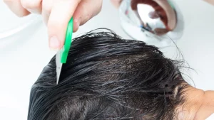 Adult hand drawing a metal fine-tooth lice comb through a small section of a child's conditioned light-brown hair in soft natural daylight, with a clean white towel folded on the wooden counter behind.