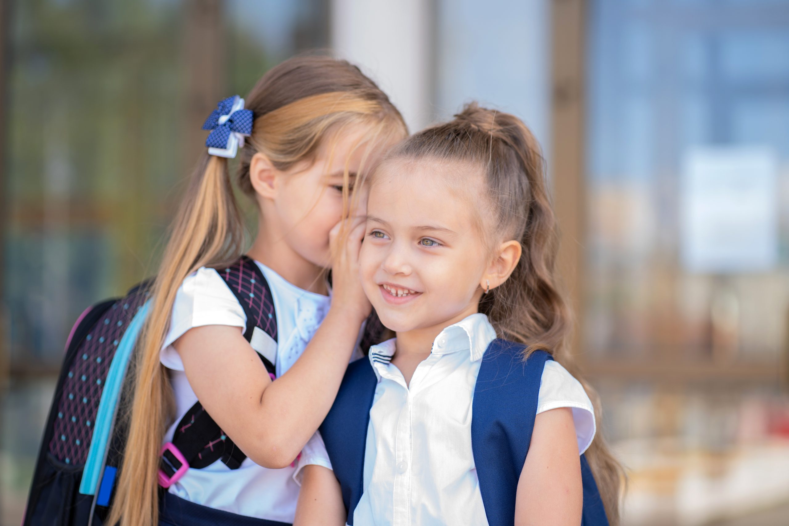 Two schoolgirls in school uniforms standing head-to-head while one whispers in the other ear, illustrating the close direct contact that actually spreads head lice between kids
