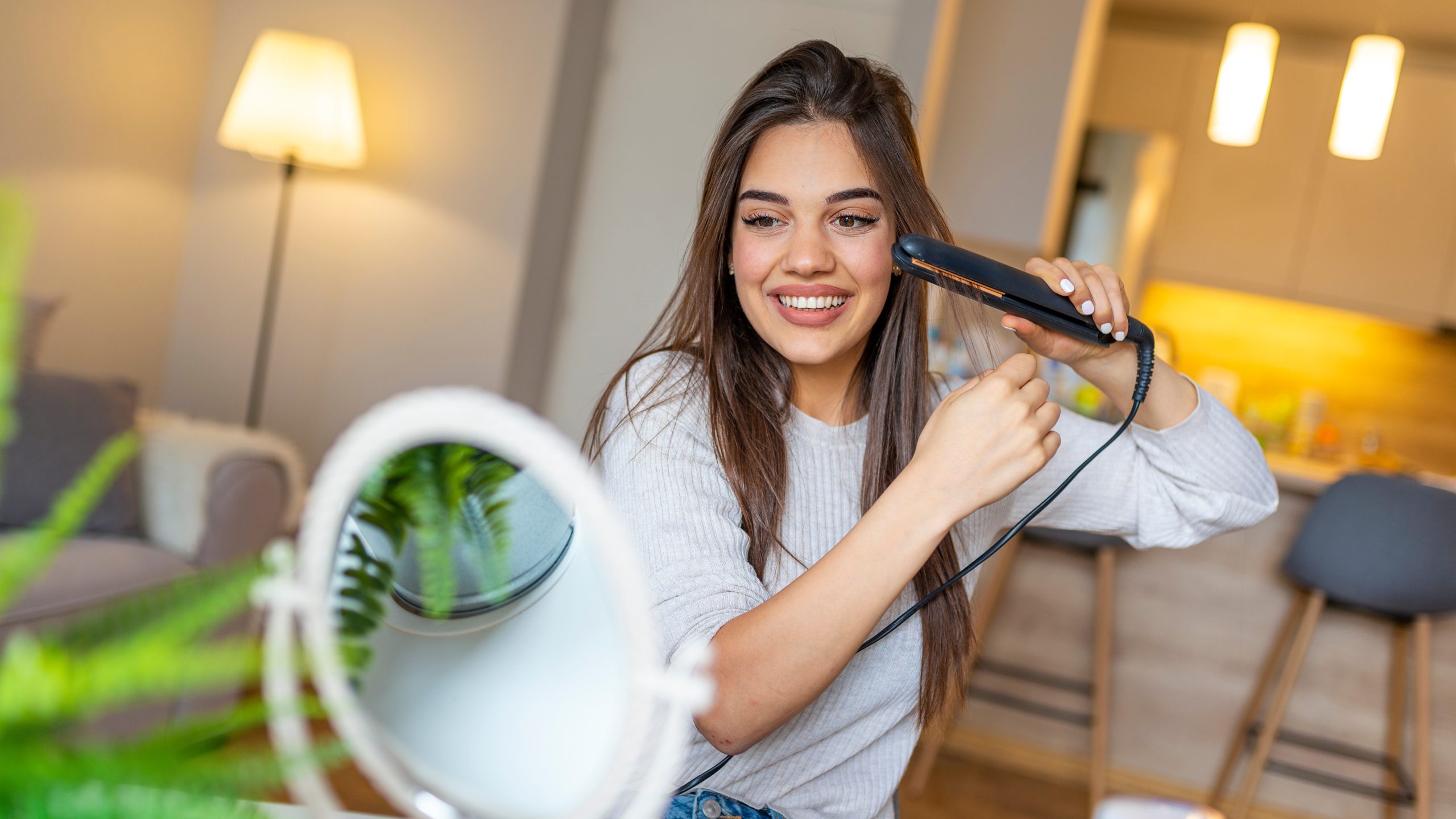 A young person uses a hot flat iron hair straightener at a bathroom mirror, the DIY styling tool many parents wrongly reach for to treat head lice.