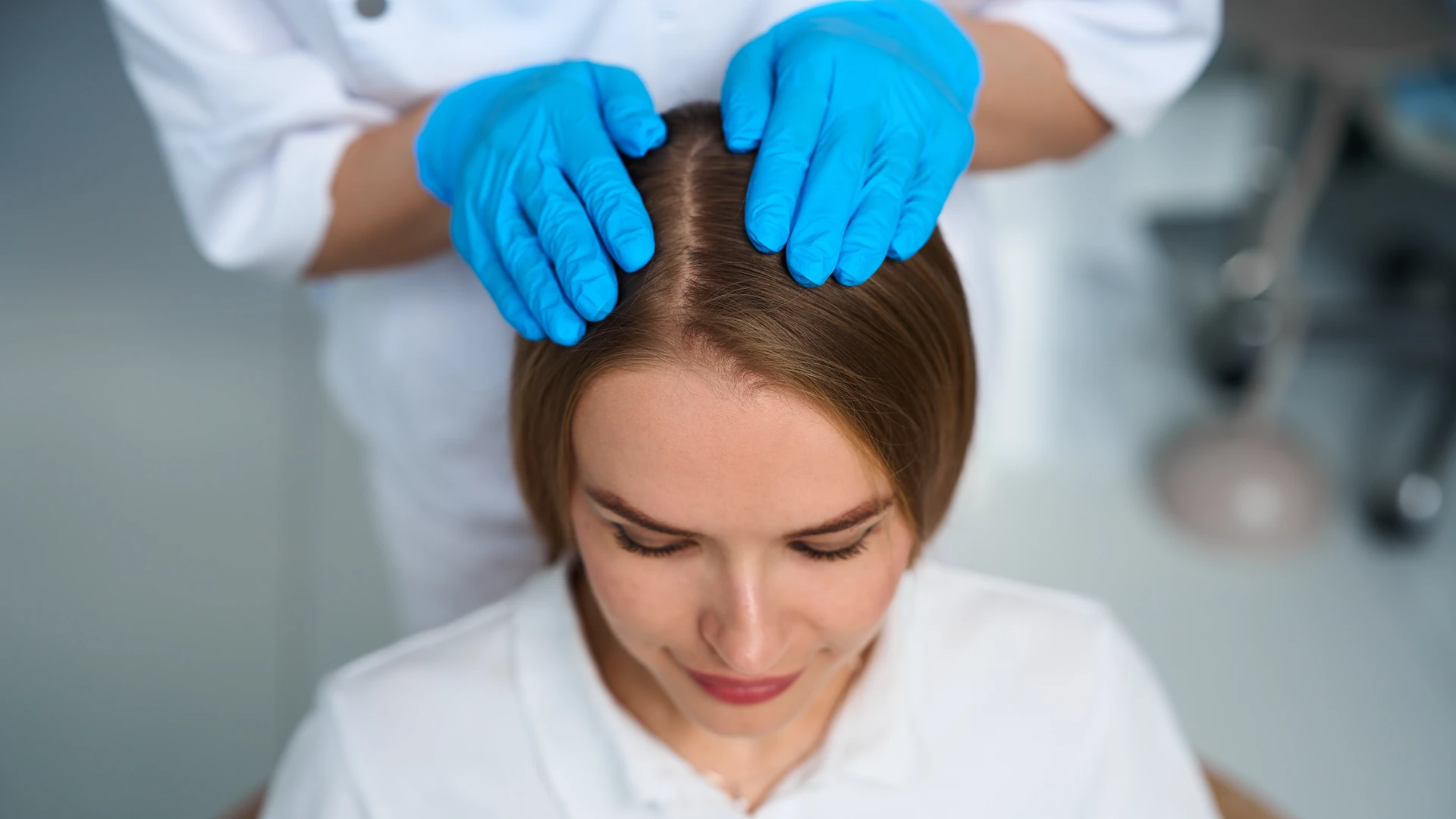 Blue-gloved hands separating brown hair along a center part.